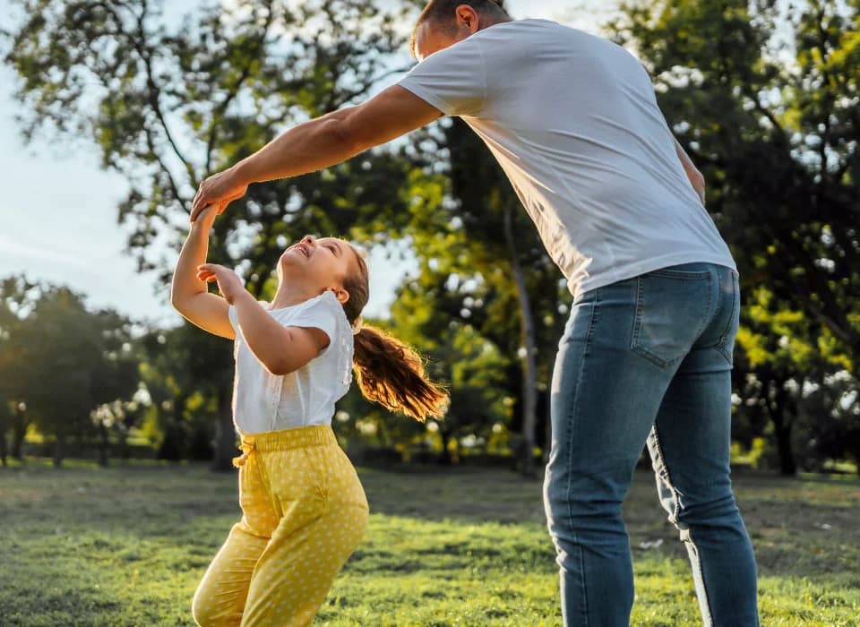 Father twirling his smiling daughter in a sunlit park, enjoying a carefree moment together.
