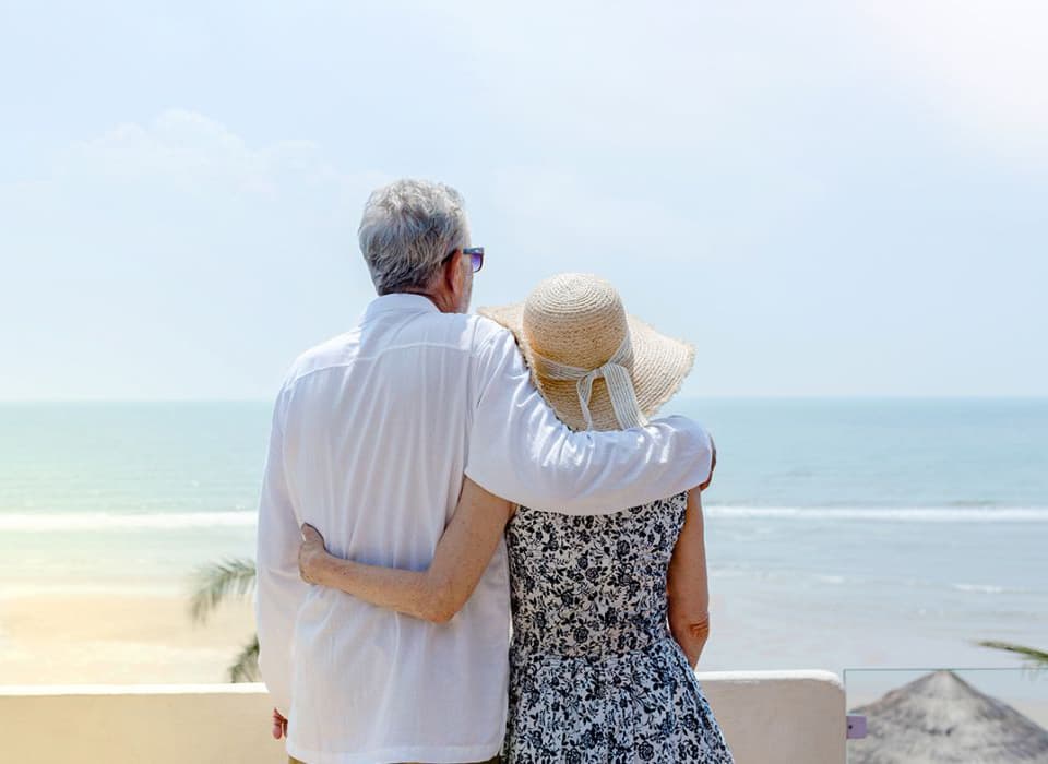 Older couple embracing and looking out over a peaceful beach, enjoying a quiet moment together.