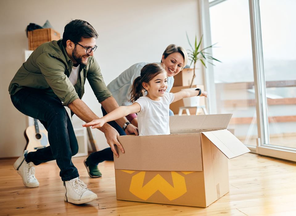 Playful family pushing a smiling child in a moving box inside their new home.