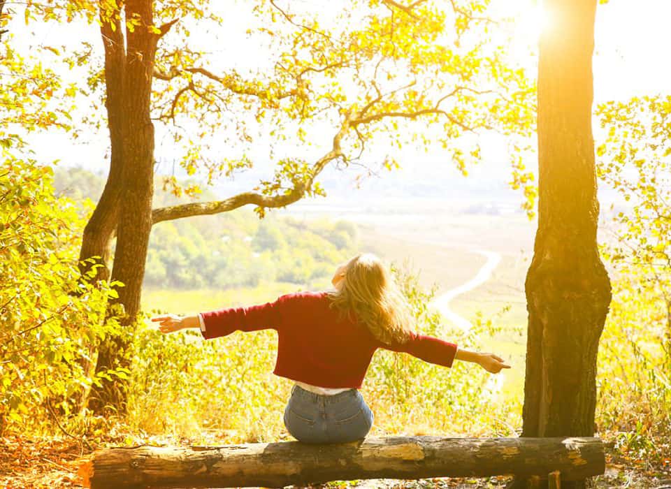 Woman sitting on a log in the forest with arms outstretched, enjoying the sunshine and nature.