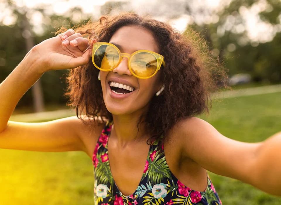 Joyful woman wearing bright yellow sunglasses and a floral dress, smiling outdoors on a sunny day.