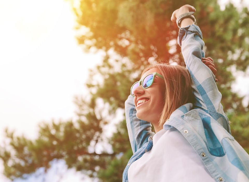 Happy young woman stretching with joy under the sun near tall trees, wearing sunglasses and a plaid shirt.