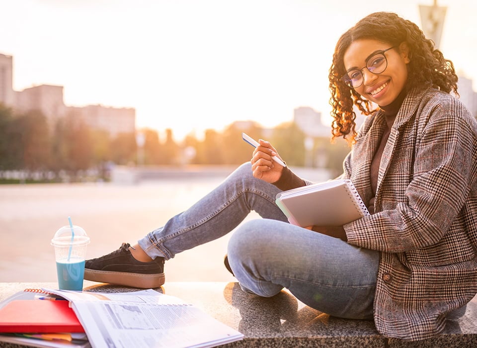 Smiling student sitting outdoors at sunset, holding a notebook and pen, with study materials and a drink on the stone ledge beside her.