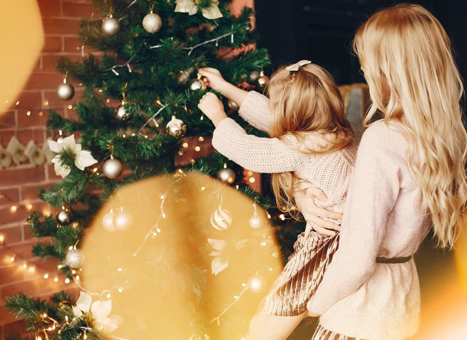 Mother helping her daughter hang ornaments on a Christmas tree, surrounded by lights and holiday decorations.