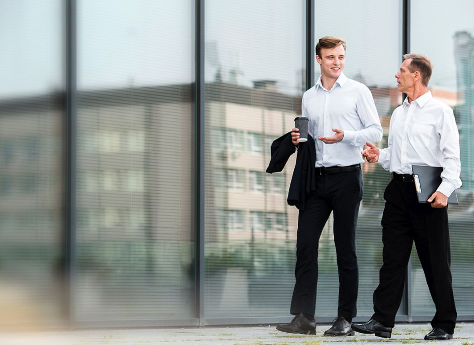 Two professionally dressed men walking and conversing outside a modern glass office building, one holding a coffee and the other a tablet.