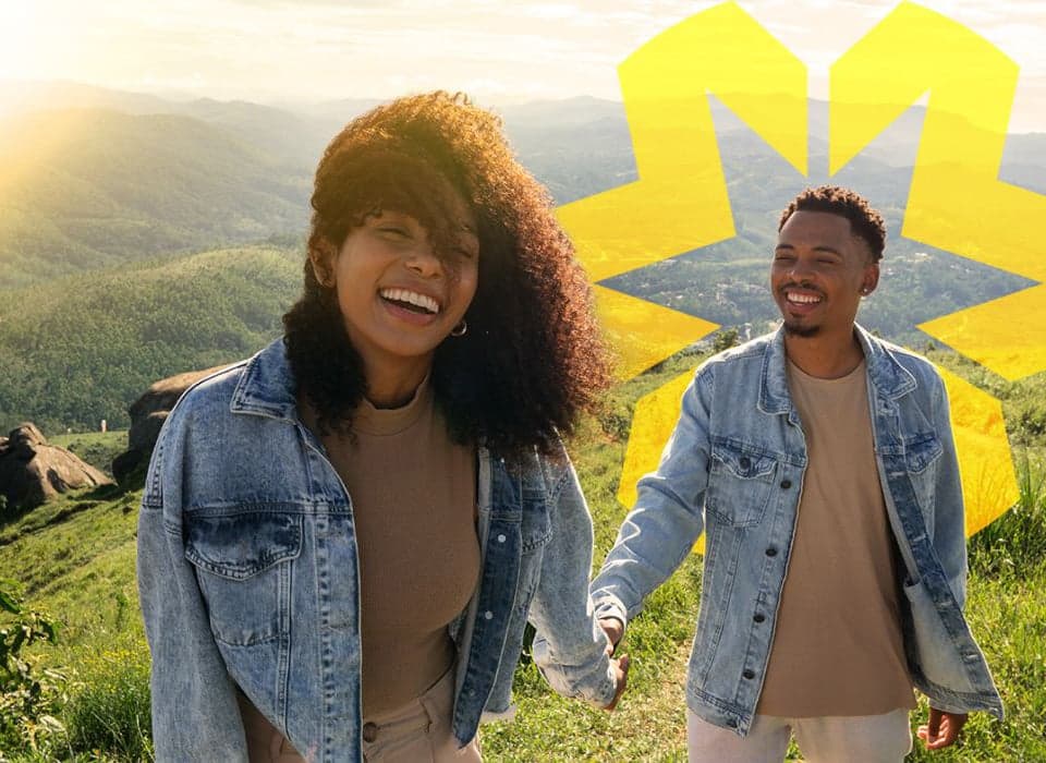 Smiling couple holding hands and hiking through a sunlit mountain landscape, dressed in denim jackets.
