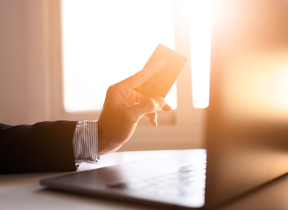 Close-up of a person’s hand holding a credit or debit card in front of a laptop, with warm sunlight streaming through a window. 