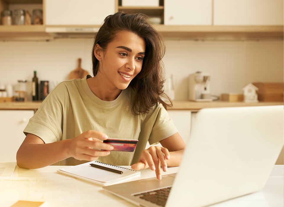 Young woman smiling while using a laptop and holding a credit card, seated at a kitchen table with a notebook. Representing secure and convenience online banking from home. 