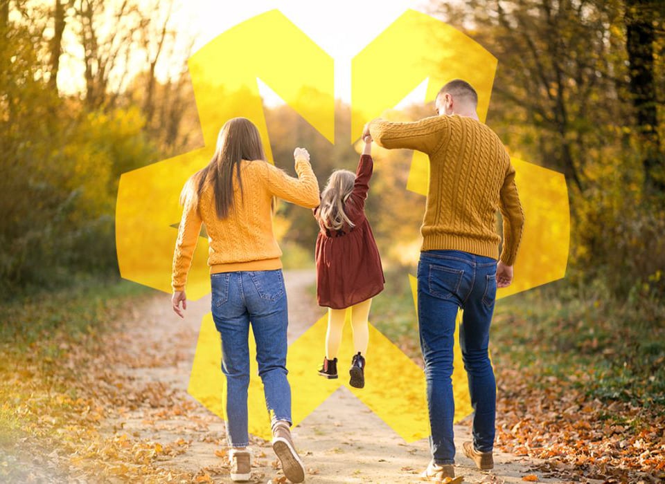 Parents swinging their young daughter while walking through a sunlit forest path in fall.