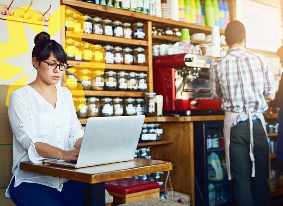 Woman working on a laptop in a cozy coffee shop with baristas preparing drinks in the background.