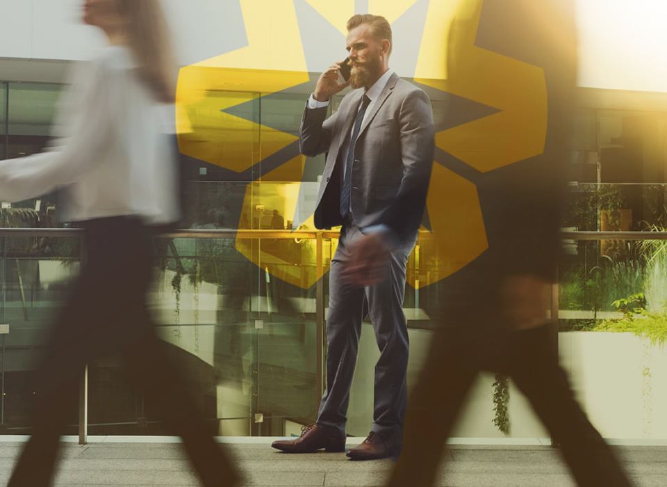 Businessman in a suit talking on his phone while others walk past in a busy office plaza.