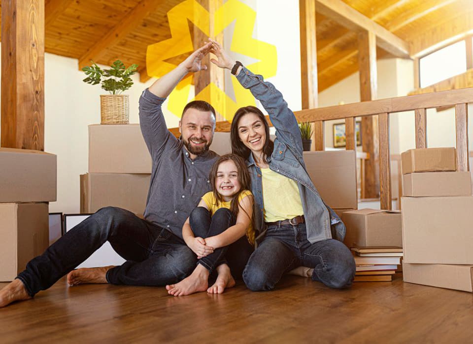 Happy family sitting on the floor of their new home surrounded by moving boxes, forming a roof shape with their arms.